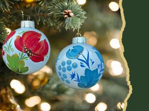 Two round Christmas tree ornaments with red and blue flower decoration hanging on a pine bough, with lights in the background