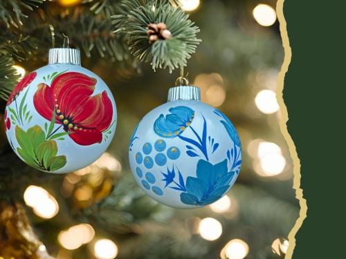 Two round Christmas tree ornaments with red and blue flower decoration hanging on a pine bough, with lights in the background