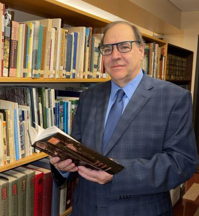 A middle-aged man with prominent dark-framed glasses in a jacket standing wth an open book in front of a booksehf