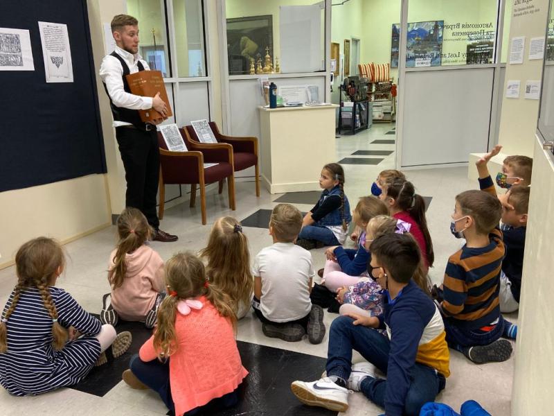 A young man standing in a gallery while holding a large book and speaking to a group of young, school-aged children who are sitting on the floor.