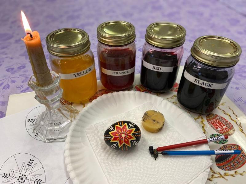 Completed pysanka, drawing tools, and block of beeswax on a paper plate, behind which are jars of colorful dyes and a lit candle.