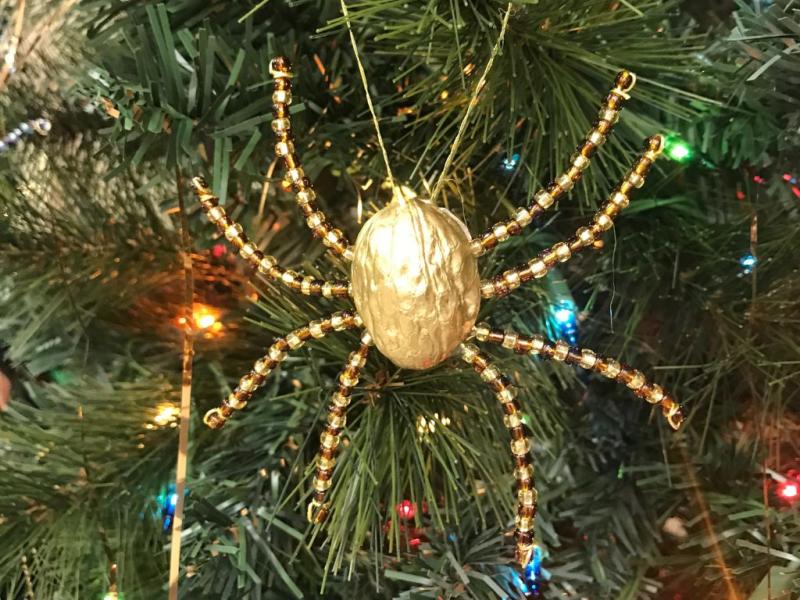 Gold painted walnut shell and gold beads in the form of a spider hanging on a pine bough with Christmas lights in the background